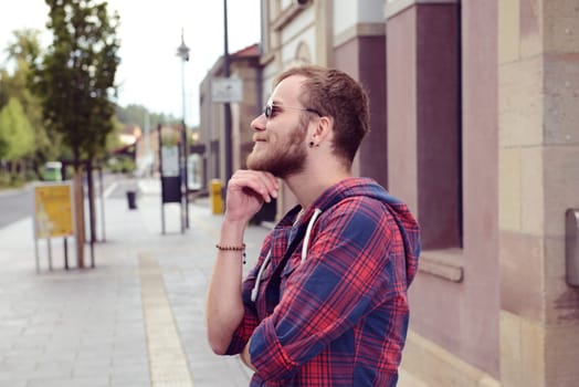young bearded man with sunglasses in checkered shirt standing in front of city background