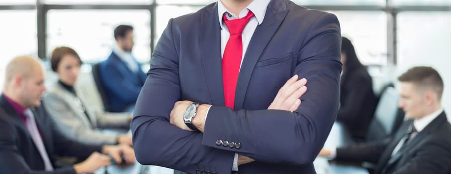 Successful team leader and business owner proudly standing with crossed arms with  coworkers working in office in background. Business and entrepreneurship concept.