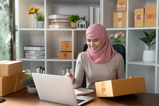 A cheerful Muslim woman in a hijab engages in her online business, managing inventory and customer orders with a laptop and tablet.