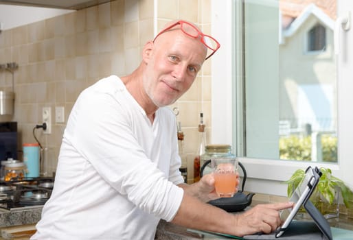 a senior man in kitchen using digital tablet and drink fruit juice