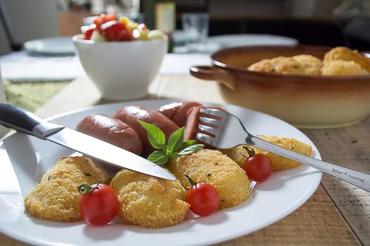 Pork sausages and breaded potatoes fried in a dish and decorated with cherry tomato. Fork and knife next to dish