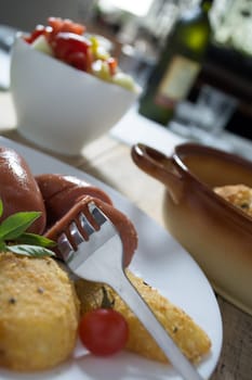 Pork sausages and breaded potatoes fried in a dish and decorated with cherry tomato. Fork and knife next to dish