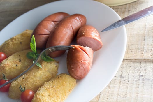 Pork sausages and breaded potatoes fried in a dish and decorated with cherry tomato.  Fork and knife placed next to dish on wooden table