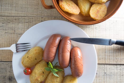 Pork sausages and breaded potatoes fried in a dish.  Fork and knife placed next to dish on wooden table