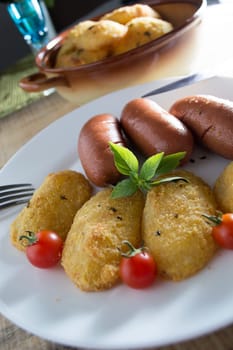 Pork sausages and breaded potatoes fried in a dish and decorated with cherry tomato.  Fork and knife placed next to dish on wooden table