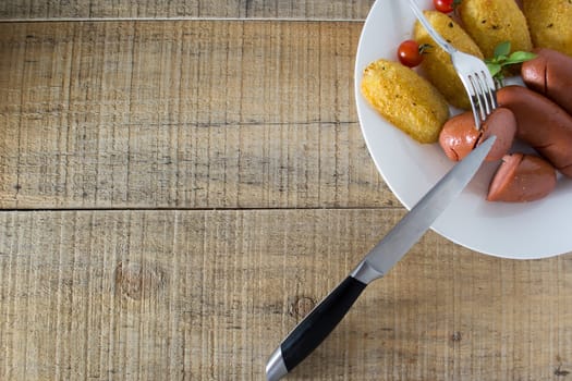 Pork sausages and breaded potatoes fried in a dish and decorated with cherry tomato. Fork and knife placed next to dish on wooden table