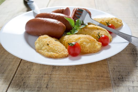 Pork sausages and breaded potatoes fried in a dish and decorated with cherry tomato.  Fork and knife placed next to dish on wooden table