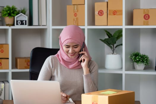 A smiling Muslim woman in a hijab speaks on the phone, overseeing her online business and packages in her home office.