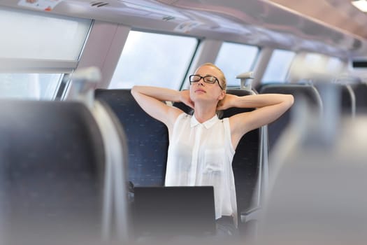 Businesswoman traveling by train, streching on her seat while working on laptop. Business travel concept.