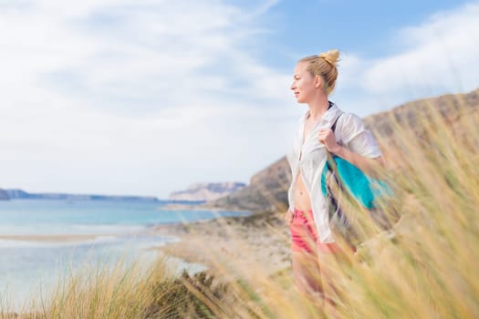 Relaxed woman on vacations in white loose shirt carrying beach bag and towel, enjoying beautiful coastline view of white sandy lagoon at Balos beach, Greece.