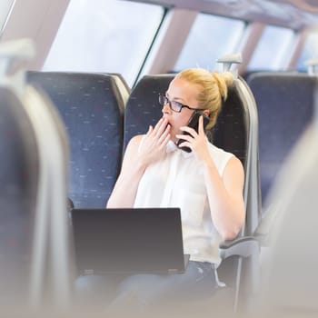 Businesswoman traveling by train, yawning while talking on cellphone and working on laptop. Long and tiresome business travel concept.