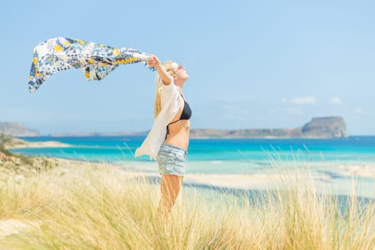 Relaxed woman, arms raised, holding colorful scarf, enjoying sun, freedom and life at beautiful beach. Young lady feeling free, relaxed and happy. Concept of vacations, freedom, joy and well being.