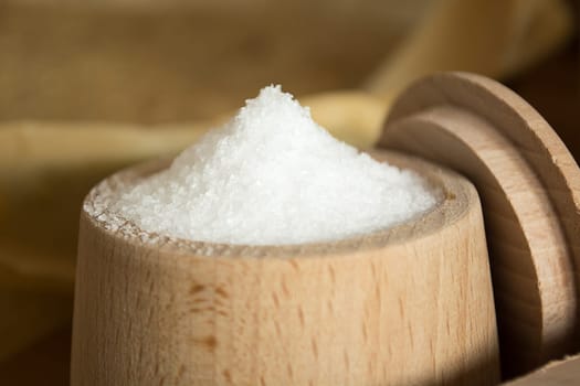 Close up of small wooden cup full of salt sitting on the kitchen table. Other kitchen wares blurred in the background