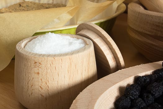 Close up of small wooden cup full of salt sitting on the kitchen table. Other kitchen wares blurred in the background