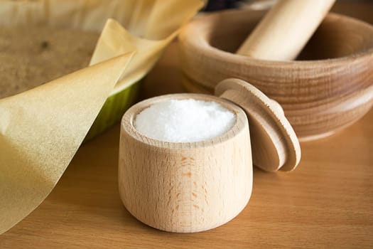 Close up of small wooden cup full of salt sitting on the kitchen table. Other kitchen wares blurred in the background