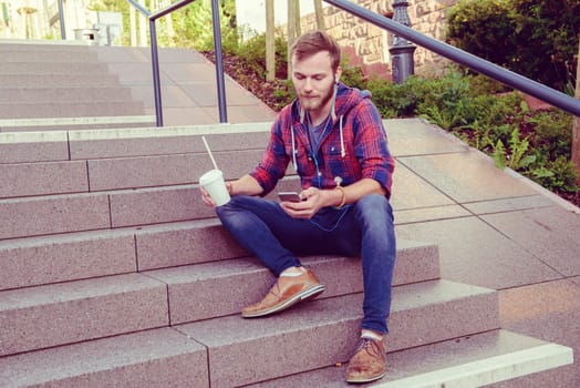 young man with soft drink sitting on stairs and using his smartphone