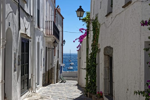 View through an alley of the Atlstadt in Cadaques in the background the sea with sailboat in the foreground the facades of the historic old houses overgrown with bougainvillea.