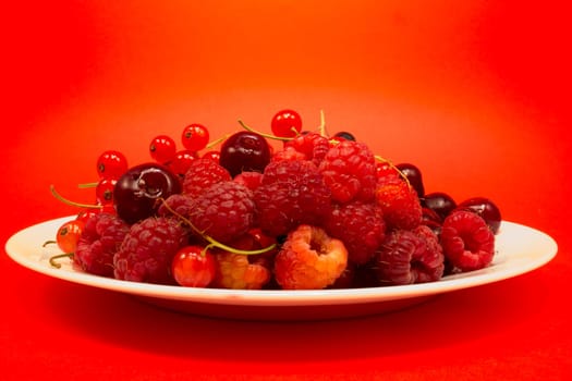 White plate with cherries, raspberries and red curant on red background