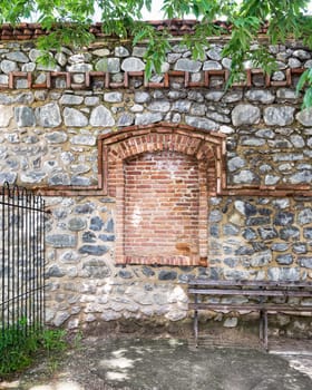 A rustic stone wall features a unique brick archway, surrounded by greenery. A simple wooden bench invites visitors to relax in this serene outdoor setting