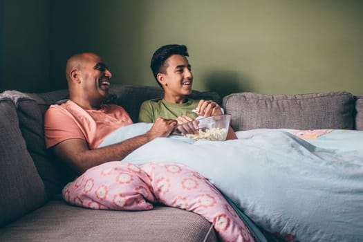 Father and son watching a film together. They are curled up on the sofa with popcorn and are laughing.