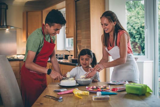 Mother decorating a cake with her son and daughter in the kitchen of their home. They are covering the cake with icing and have sweets to decorate.