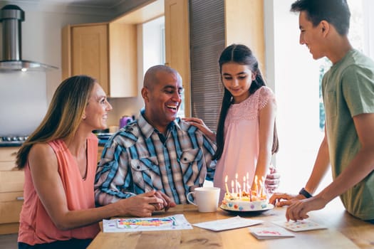 Family of four celebrating the fathers birthday with a cake that the children made. He is laughing as they sing before he blows out the candles.