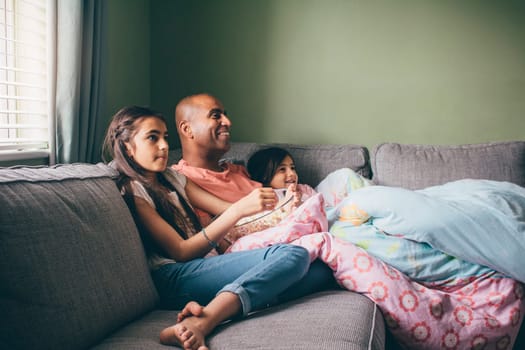 Father curled up on the sofa with his two daughters. They are watching a movie with popcorn.