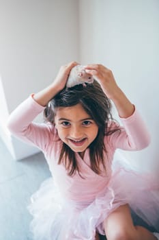 Little girl sitting on the floor in her ballet costume. She has her pet hamster on her head and is looking up to the camera with a smile on her face.