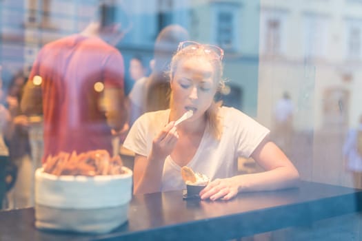 Young pretty woman eating icecream in gelateria. Through the window view.