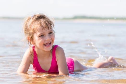 Five-year girl lies in water in the shallows of the river, laughing and looking to the left