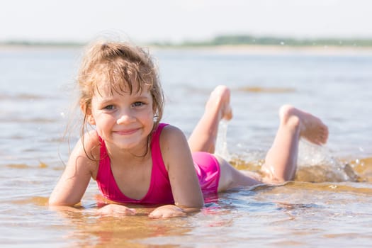 Five-year girl lies in water in the shallows of the river and looking at the frame