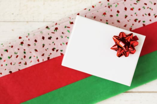Small white gift box with red bow on colorful red and green tissue paper.