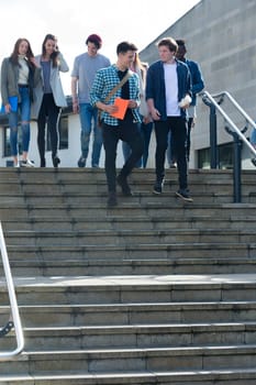 A group of students laugh and smile as they leave one of their lectures. They are walking down some steps carrying their books, bags and digital tablets.