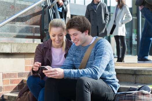A happy young couple smile as they sit on some steps in the city looking at a mobile phone. There is a group of other students out of focus in the background behind them.