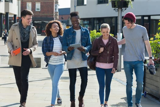 A happy group of friends smile as they walk through the city together. They are carrying their bags and digital tablets on a break from university.