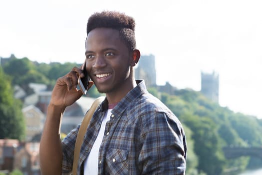 A young man uses his mobile phone outdoors next to a river. He is talking to someone. A cathederal can be seen in the background.