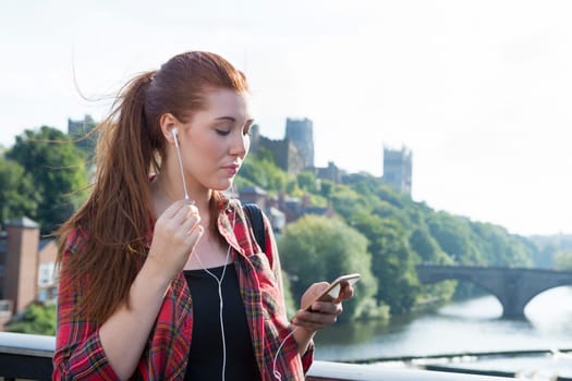 A happy young woman smiles as she looks down at her phone listening to her music by the river. She has her hair pulled back into a ponytail with her bag over her shoulder.