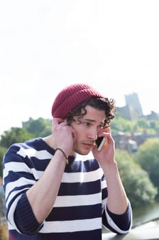 A young man uses his mobile phone outdoors. He is holding his phone to one ear and his hand to his other try and hear the person better.