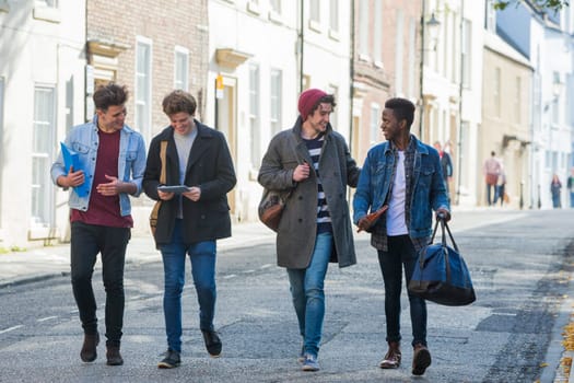 A happy group of male students smile and laugh as they walk through the city together. They are carrying their bags and books.