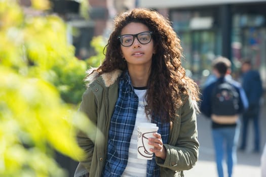 A happy female student looks at the camera as she walks through the city. She is carrying her hot drink and is wrapped up warm for the colder weather.