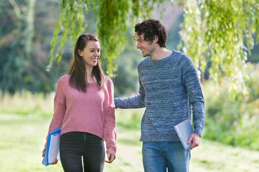 A happy young couple smile at each other as they walk through a country park together. They are carrying their school books and the man is holding his grilfriend's arm.