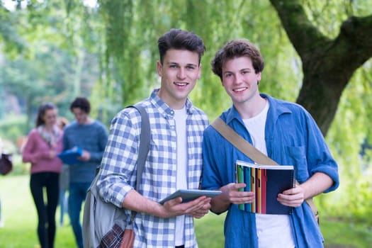Two happy male students smile as they relax outdoors and study. They are holding their bags, books and a digital tablet as they look at the camera.