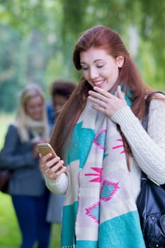 A happy yung teenager smiles as she looks at her phone. She is standing outdoors wrapped up warm. A group of girls are in the background out of focus.