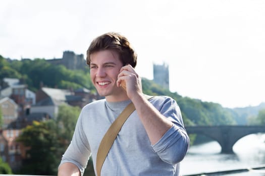 A young man uses his mobile phone outdoors next to a river. He is talking to someone. A cathederal can be seen in the background.