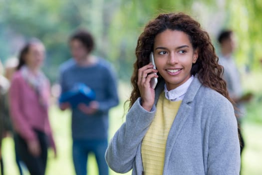 A happy young student smiles as she uses her mobile phone outdoors. She is wrapped up in a thick coat. A group of students are out of focus in the background.