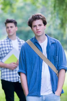 A happy teenage student smiles at the camera as he stands outdoors with his hands in his pockets. His friend is behind him out of focus. He has his bag across his shoulder.