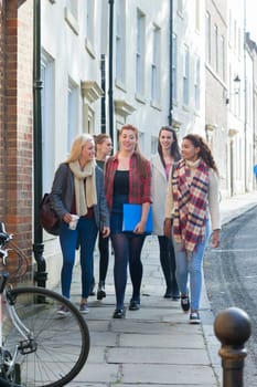 A group of young females smile as they walk through a city together on a break from studying. They are carrying their bags and books along withe them.