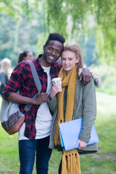 A happy young couple smiles as they relax outdoors with their friends. They are on a break from studying standing in an embrace and the girl is holding her books and a hot drink.