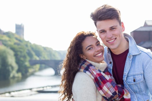 A happy young couple smile at the camera as they relax outdoors by the river. They are wrapped up warm for the winter.