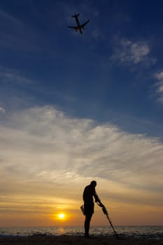 a treasure hunter with Metal detector on sunset on the beach with a plan in the sky